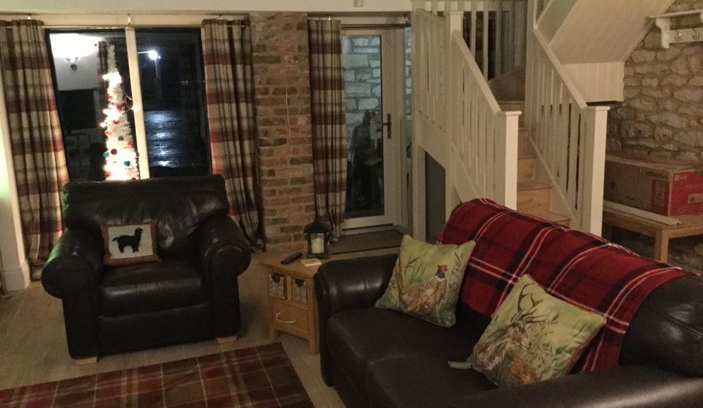 Living room of holiday cottage showing brown leather 3-seater settee and armchair with L-shaped staircase to the background on the right and patio doors to background on the left