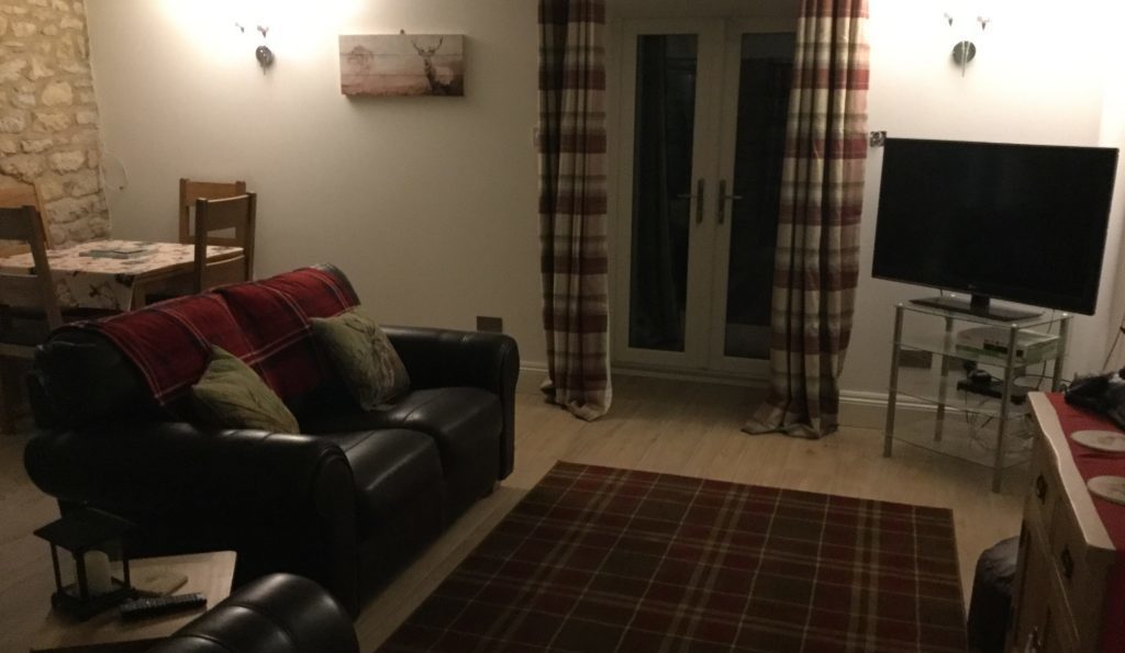Picture of holiday cottage living room showing dining table on the left, 3 seater brown leather sofa, TV to the right, overlooking brown leather chair