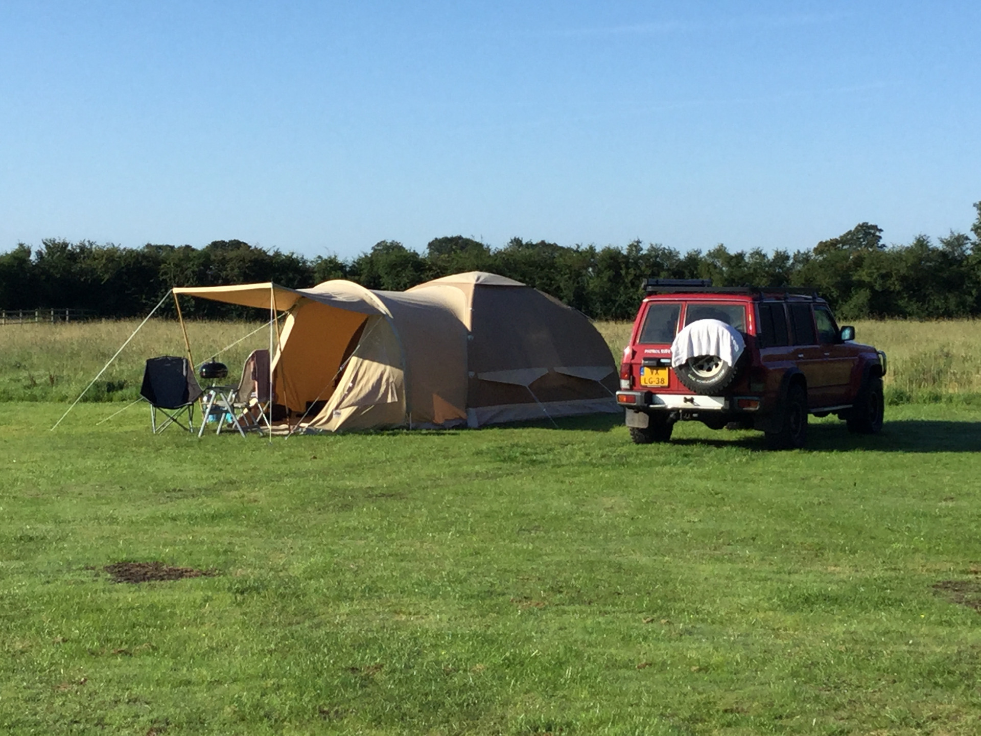 Beige tent with awning pitched next to a red 4x4 vehicle, on lush green grass and with blue sky